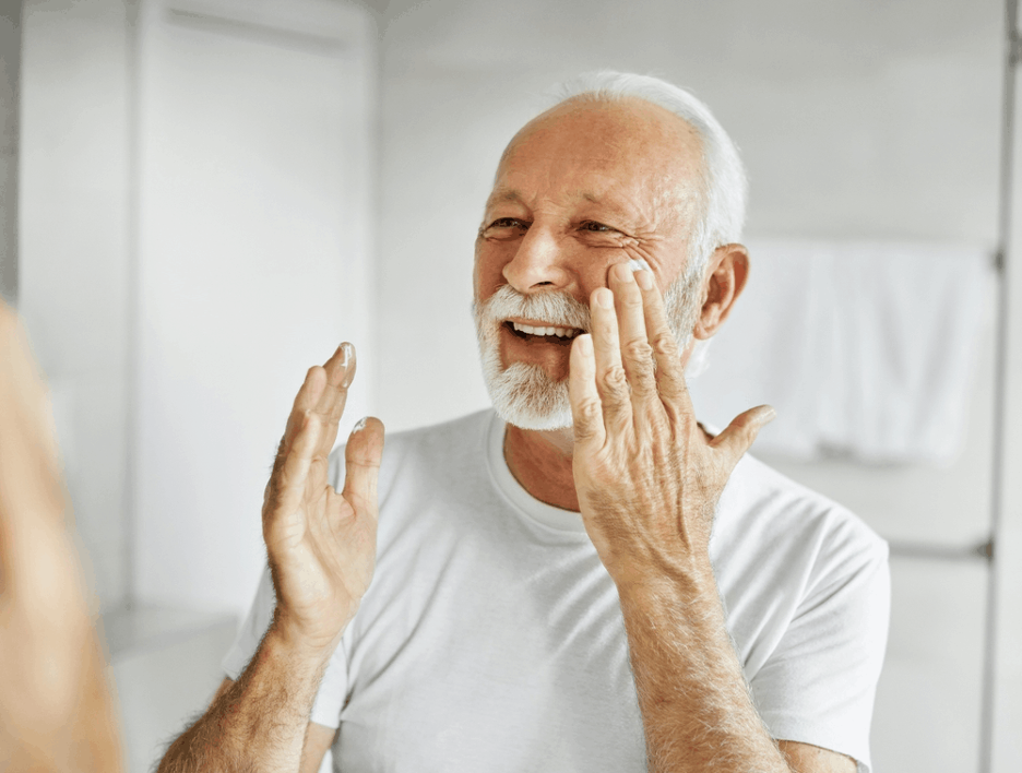 Older man with white beard applying moisturizer to his face in a bright bathroom